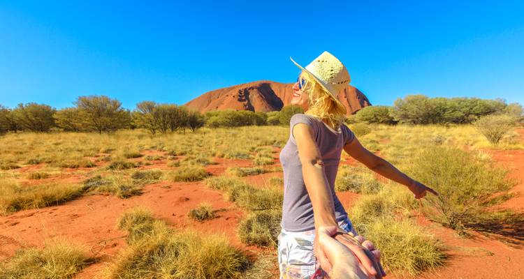 Viajero sonriente guiando al espectador hacia Uluru a través de matorrales del desierto rojo bajo un cielo azul despejado.