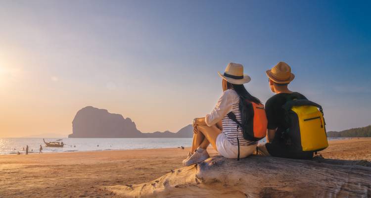 Pareja joven con mochilas sentada en un tronco de madera flotante, viendo el atardecer sobre una playa tropical tranquila y una isla kárstica distante.