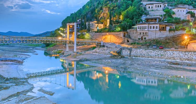 Evening lights reflect in a calm river beside historic stone houses and a pedestrian bridge in Berat.