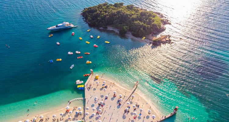 Vibrant aerial of a sandy tongue beach, colorful kayaks and a small wooded island in dazzling turquoise water.