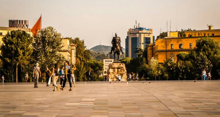 A broad plaza with locals and tourists faces an equestrian statue and modern buildings in Tirana.