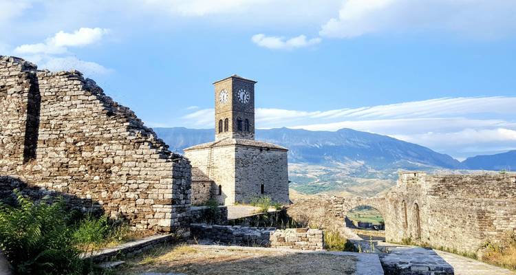 Ruins of a stone clock tower stand on a ridge with sweeping mountain vistas under blue skies.