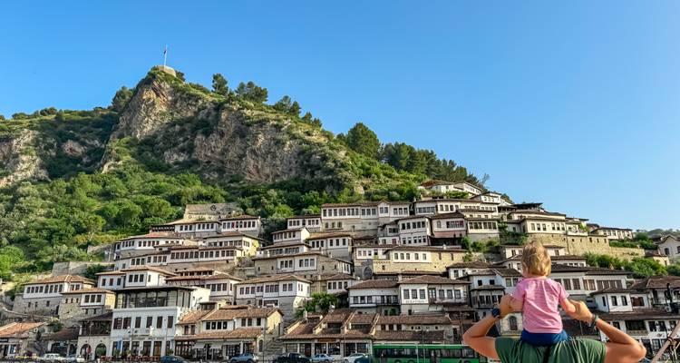 A parent lifts a child on shoulders before the terraced Ottoman houses of Berat climbing a green hillside.
