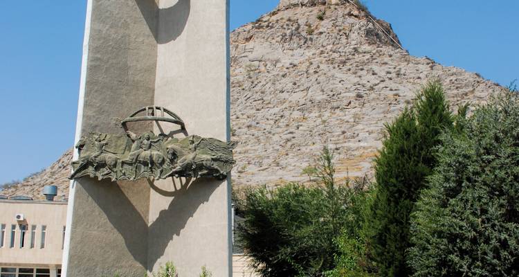Sovjet-tijdperk monument met reliëfsculptuur geplaatst voor een rotsachtige heuvel onder een heldere blauwe hemel.