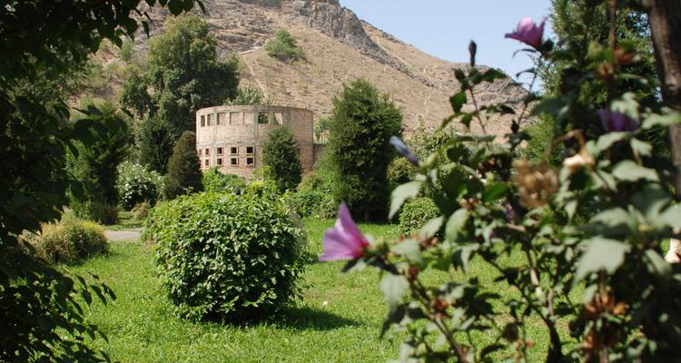 Cirkelvormige stenen ruïne gedeeltelijk verborgen tussen weelderig groen met berghelling erachter.