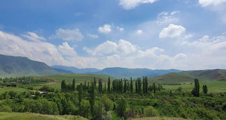 Brede pastorale vallei met rijen populieren en verre glooiende bergen onder verspreide wolken.