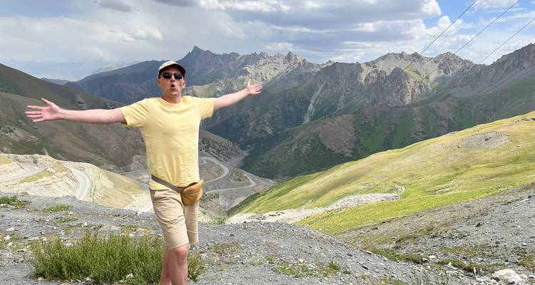 Jonge man in casual kleding poseert met uitgestrekte armen op een hoge bergpas met uitzicht op kronkelende wegen en ruige bergtoppen.