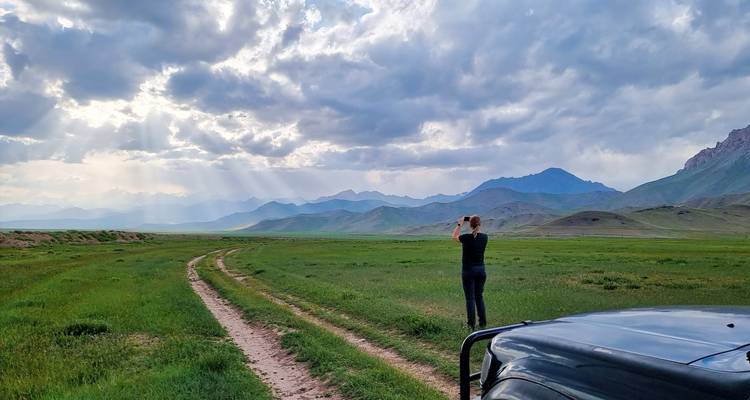 Persoon met camera staat op een zandpad over een uitgestrekte groene vallei omlijst door dramatische bergen en door de zon beschenen wolken.