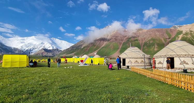 Kleurrijk joerten- en tentenkamp op weelderig grasland onder besneeuwde bergen onder een heldere blauwe hemel.