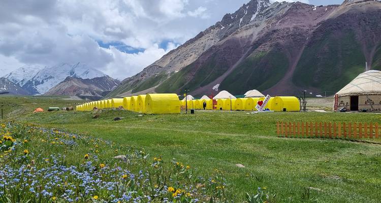 Lange rij heldergele expeditietenten op bergweide met wilde bloemen, besneeuwde toppen en dramatische bergkammen op de achtergrond.