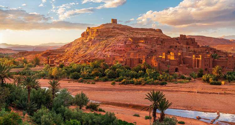 Adobe hilltop ksar of Ait Ben Haddou glowing in warm golden hour light with palm-lined riverbed below.