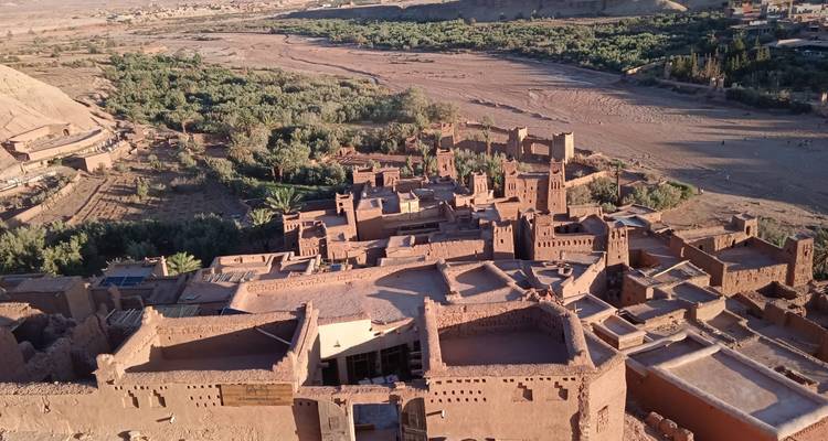 Aerial view of fortified adobe settlement and surrounding oasis along a dry riverbed in the desert.
