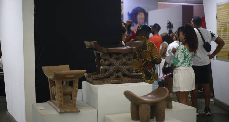 Exposition intérieure de musée de tabourets en bois sculptés avec des visiteurs examinant les pièces exposées.