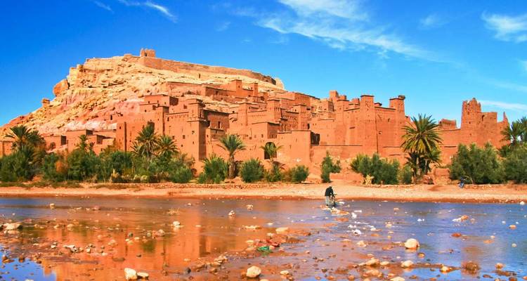 Daytime view of fortified Ait Ben Haddou with shallow river in foreground where a lone figure crosses.
