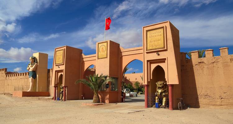 Entrance to Atlas Film Studios with desert-style walls, statues and Moroccan flag under blue sky.