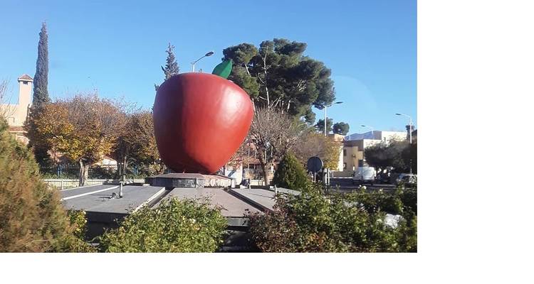 Large red apple sculpture standing on a roundabout surrounded by greenery under clear blue sky.
