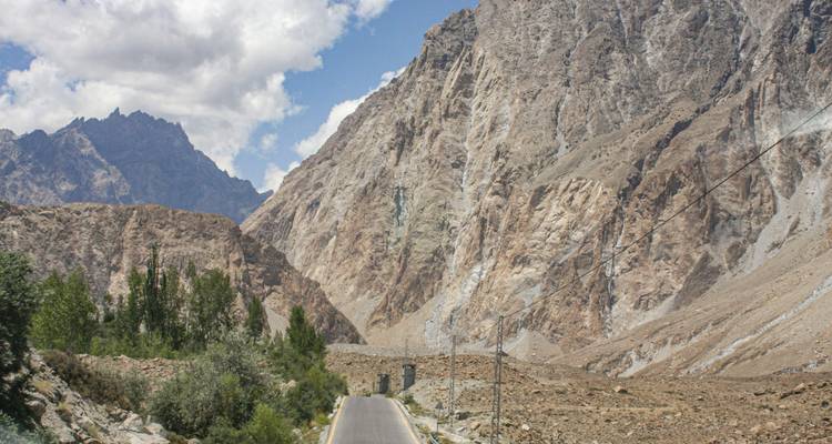 Narrow asphalt highway cutting through towering rocky mountains under partly cloudy sky