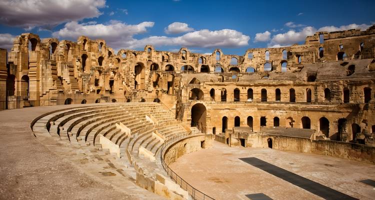 Gut erhaltenes römisches Amphitheater von El Jem mit weitläufigen Steinsitzreihen unter einem blauen Himmel mit Wolken.