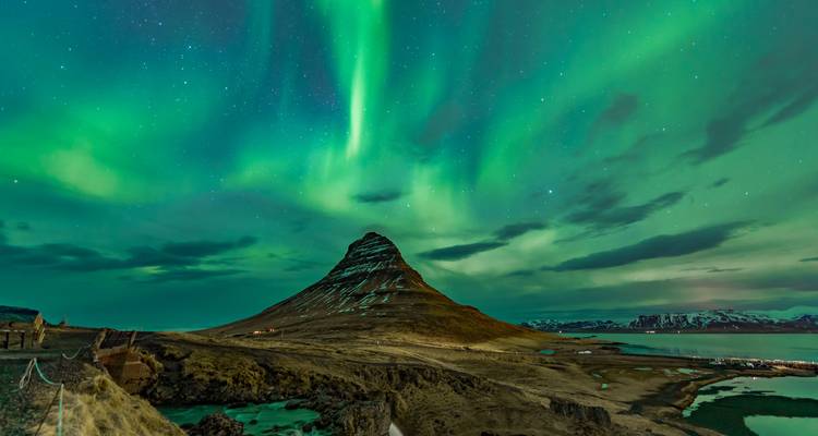Des aurores boréales d'un vert éclatant tourbillonnent au-dessus de la montagne conique Kirkjufell près d'un fjord immobile sous un ciel nocturne étoilé.