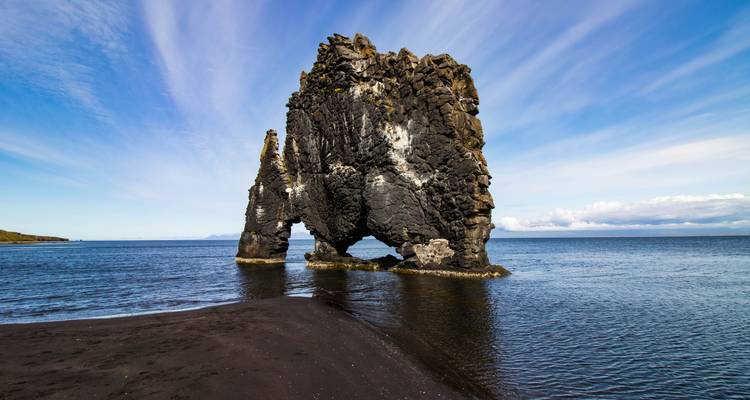 La pile de mer en basalte noir de Hvítserkur s'élève des eaux bleues calmes et du rivage de sable noir sous un ciel strié.