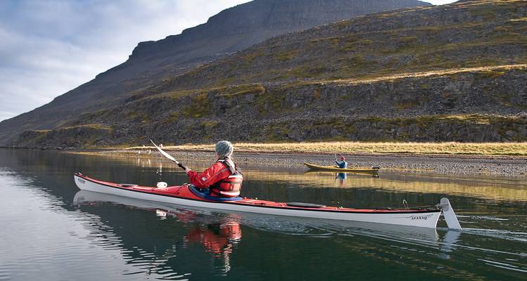 Kayaker in red paddling alongside rugged cliffs of an Icelandic fjord
