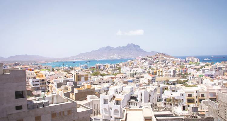 Vista panorámica de la ciudad de Mindelo con edificios coloridos de poca altura, bahía turquesa y picos escarpados al fondo.