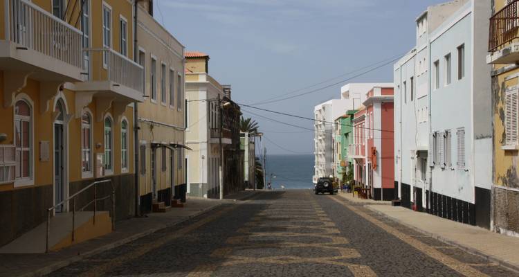 Rue pavée tranquille et colorée descendant vers la mer entre des bâtiments coloniaux pastel.