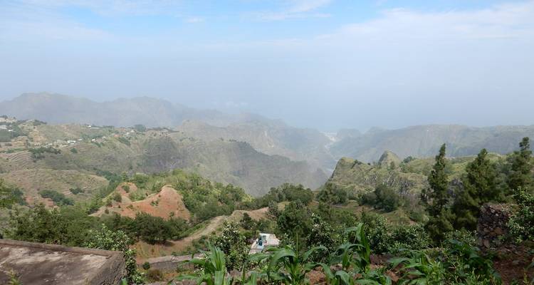 Panorama montagneux brumeux vu depuis une colline agricole avec des plants de maïs au premier plan.