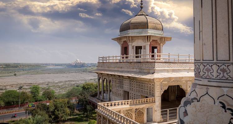 Eleganter Mogul-Pavillon der Festung Agra mit Blick auf eine weite sandige Flussebene mit dem fernen Taj Mahal unter dramatischen Wolken.
