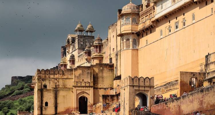 Massive ockerfarbene Mauern und Bastionen des Amber Fort erheben sich gegen einen stürmischen Himmel mit einigen Touristen und Elefanten darunter.