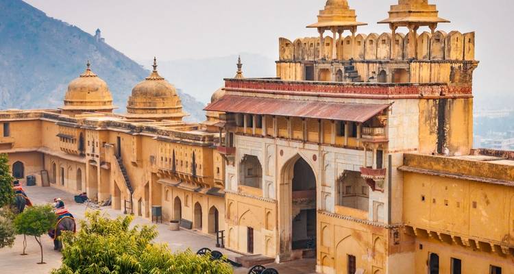 Weitläufiger Blick über die Festungsmauern von Amber Fort mit Kuppeln, Elefanten mit Reitern und den dunstig daliegenden Aravalli-Bergen dahinter.
