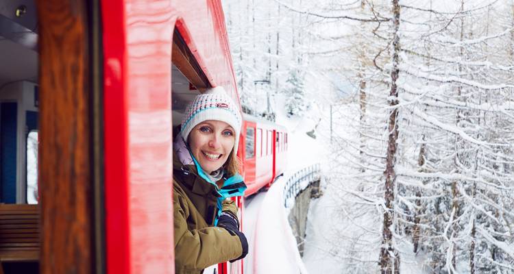 Un passager souriant se penche par la fenêtre d'un train de montagne rouge vif qui serpente à travers un paysage forestier enneigé.