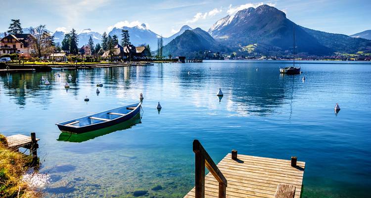 Le lac d'Annecy cristallin reflète les montagnes aux sommets enneigés et un ciel lumineux, avec de petits bateaux amarrés près d'une jetée en bois.
