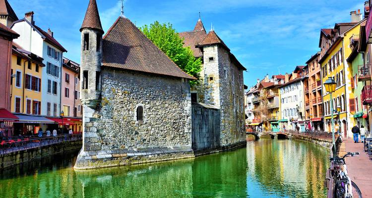Vue de jour du Palais de l'Isle en pierre encadré par des façades colorées le long du canal émeraude d'Annecy.