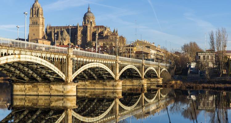 Historische stenen brug gespiegeld in kalm water leidend naar Salamanca's kathedraalcomplex gebaad in warm licht.