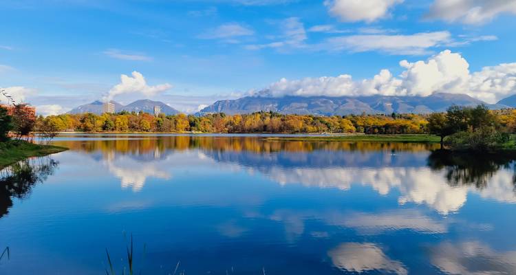 Lac calme reflétant les arbres d'automne et les montagnes lointaines sous un ciel bleu dégagé.
