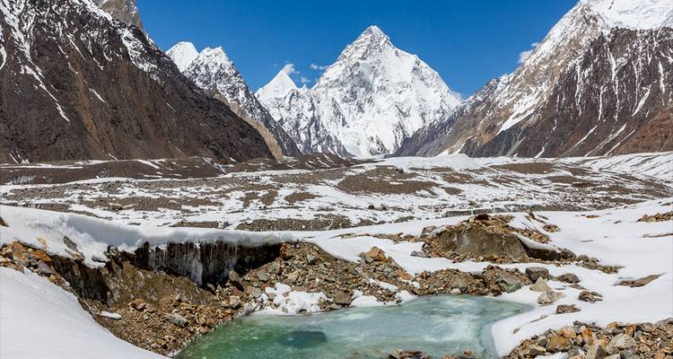 Schneebedeckte Himalaya-Gipfel erheben sich hinter einem gefrorenen Bach und einem türkisfarbenen Schmelzwassertümpel in einem Hochgebirgstal.