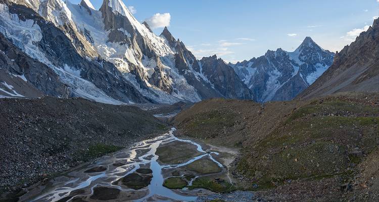 Ein gewundener Schmelzwasserfluss schlängelt sich durch ein felsiges Tal, das von hoch aufragenden eisbedeckten Gipfeln eingerahmt wird.