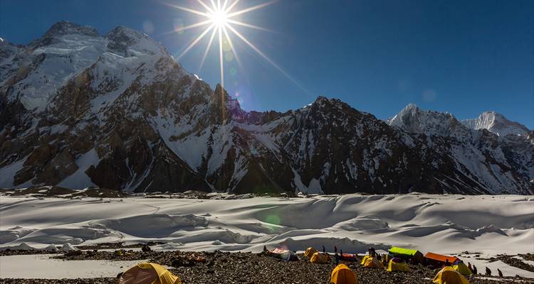 Heller Sonnenschein über schneebedeckten Gipfeln beleuchtet ein Basislager aus gelben Zelten auf einem weiten Gletscher.