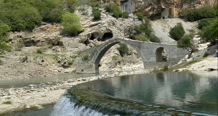 Pont historique en arc de pierre enjambant une rivière claire avec des berges rocheuses et une petite cascade.