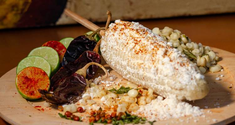 Épi de maïs de rue mexicain (elote) garni de fromage, piments, citron vert et épices sur une planche de bois.