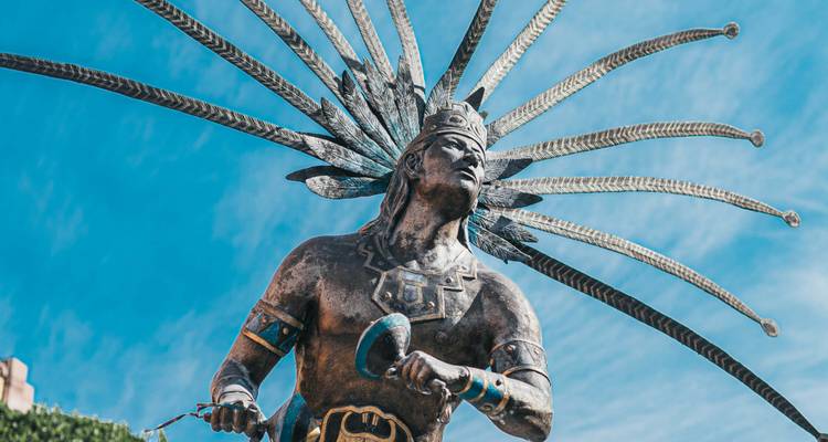 Bronzen standbeeld van een inheemse danser met een ratel en een dramatische veertooi tegen een blauwe lucht.