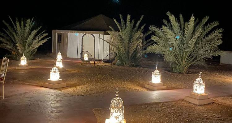 Campement du désert la nuit avec des palmiers et des lanternes lumineuses bordant un sentier menant à une tente en toile.