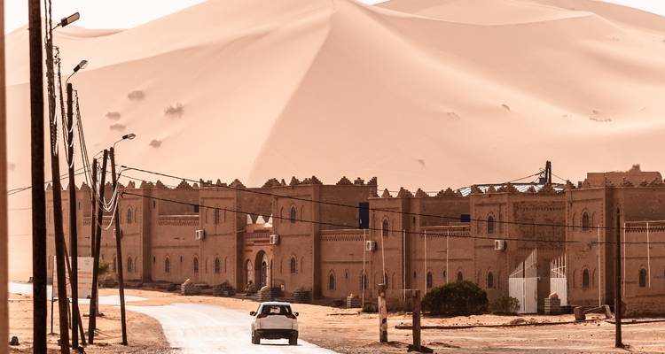 Un coche se dirige hacia un complejo de casba de adobe situado frente a una duna imponente.