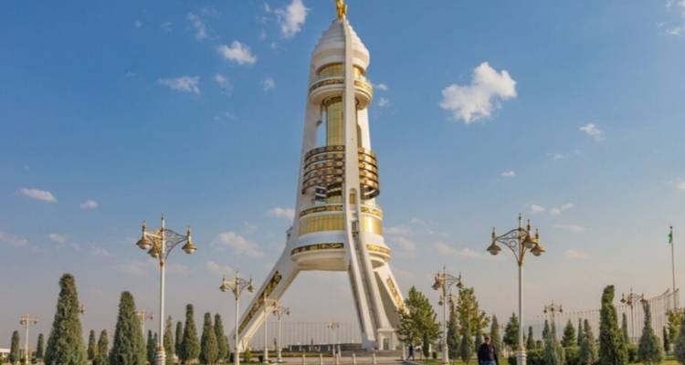 Monumento moderno alto y blanco con detalles dorados se eleva sobre jardines paisajísticos bajo un cielo azul brillante.