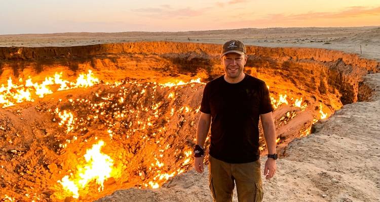 Un viajero se encuentra junto al ardiente cráter de gas de Darvaza que brilla contra el desierto al atardecer.