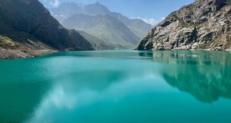 El lago alpino turquesa refleja acantilados grises escarpados y picos distantes espolvoreados de nieve bajo un cielo brillante.
