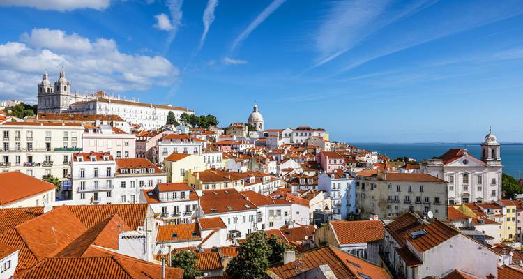 Klassiek Alfama panorama van witte gebouwen met rode daken die naar de blauwe Taag-monding afdalen onder een gestreepte hemel.