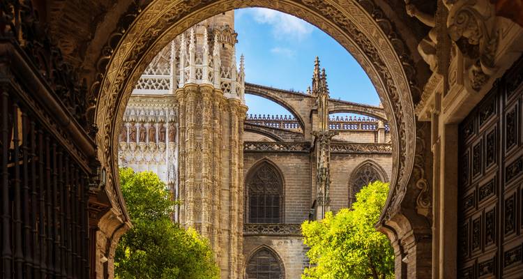 Zicht door sierlijke boog die de oprijzende steunberen van de kathedraal van Sevilla en een binnenplaats met sinaasappelbomen omlijst.