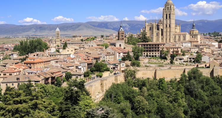 Heuveltopuitzicht op de skyline van Segovia gedomineerd door zijn majestueuze kathedraal, met bergen en blauwe lucht daarachter.